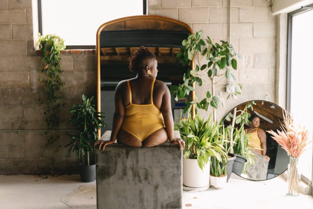 Individual boudoir session client is wearing a strappy yellow bodysuit, sitting on gray chair in front of ornate mirror and plants